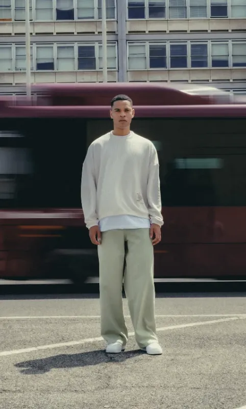Ragazzo con maglione color crema e pantaloni salvia in strada, con un autobus in movimento